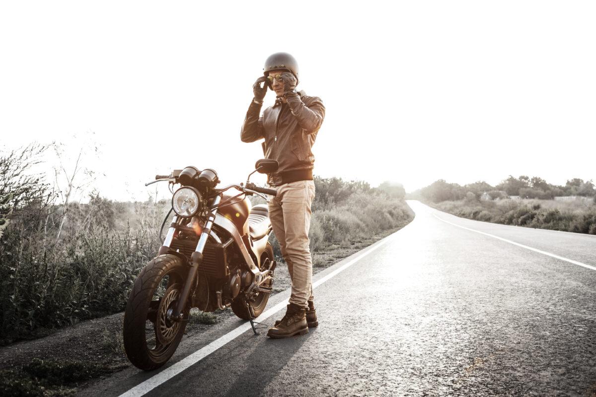 Young handsome man posing near his motorbike at countryside road. Flare sunlight background.
